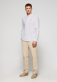 Vit långärmad skjorta med knäppning och mandarin-krage, beige byxor och ljusbruna espadrillos. Har en liten logotyp på bröstet.