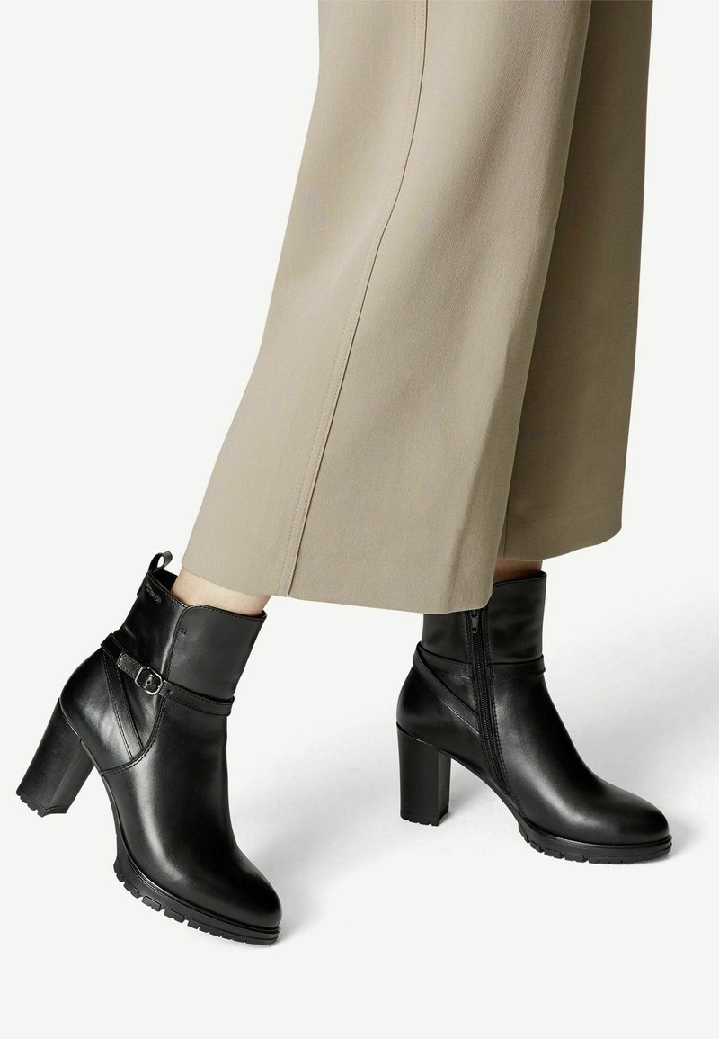 Bottes basses en cuir noir avec talons carrés hauts et détail de boucle, portées avec un pantalon beige à jambes larges sur un fond blanc uni.