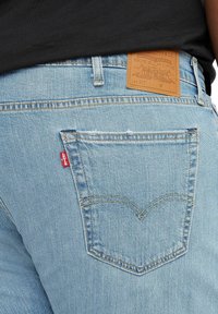 Jasnoniebieskie dżinsy z denimu mają skórzaną naszywkę na pasku z wytłoczonym tekstem, małą czerwoną metkę na tylnej kieszeni oraz standardowe przeszycia kieszeni.