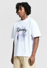 Valkoinen puuvillatakki, jossa on värikäs graafinen printti kaupunkinäkymästä ja sana "Relevity" mustalla käsinkirjoitetulla fontilla. Rento malli, jossa on lyhyet hihat.