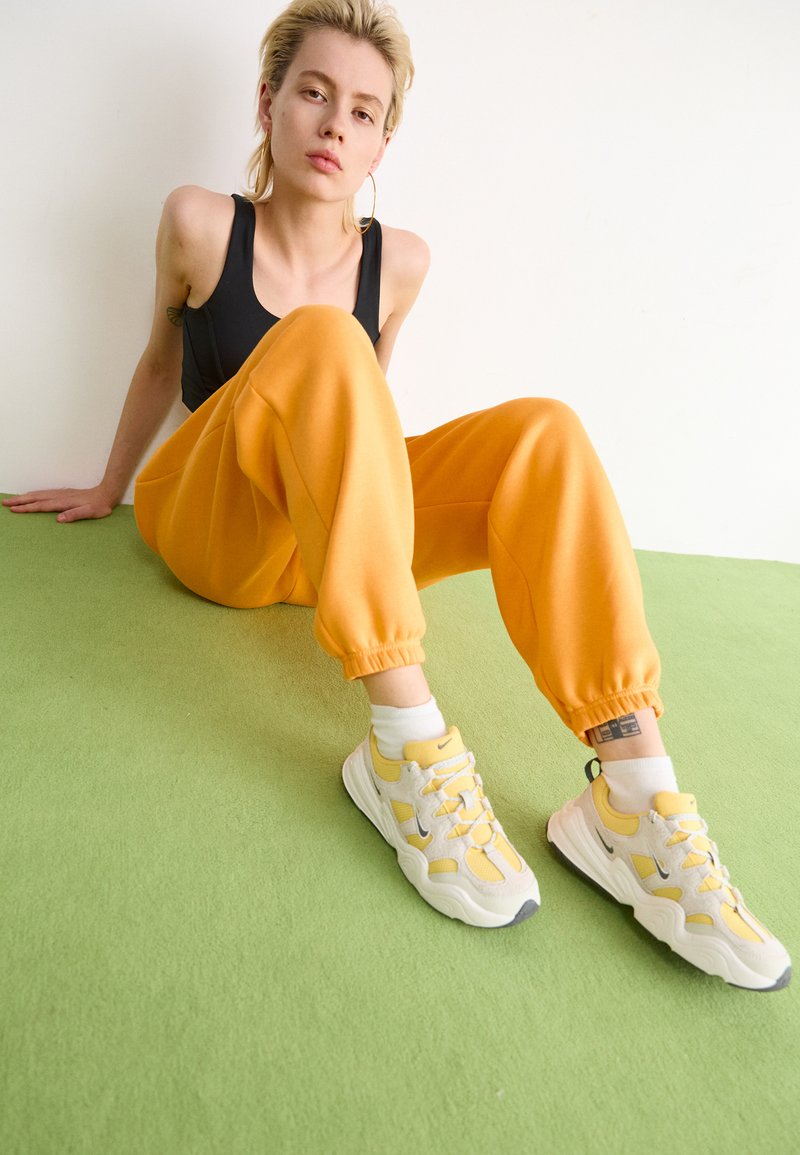 Pantalons de survêtement orange avec des poignets élastiques, associés à des baskets jaune clair avec des accents gris. Débardeur noir, assis sur une surface verte.