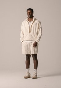 Light beige zip-up jacket with a relaxed fit, paired with matching shorts. Feet are adorned with beige sneakers and white socks.