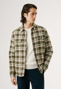 Camisa de franela a cuadros verdes con acentos en blanco, rojo y verde oscuro; presenta dos bolsillos en el pecho y un cuello clásico; llevada sobre un suéter ligero.