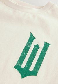 Cremefarbenes T-Shirt mit einem abgenutzten grünen Grafikdesign und spitz zulaufenden Akzenten, das den stilisierten Buchstabenformen ähnelt.