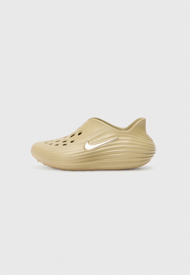 Nike slip-on skor i ljusbrun, med ribbat mönster, flera ventilationshål och en silverfärgad logotillbehör på sidan.