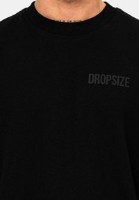 Sort bomulds t-shirt med rund hals og korte ærmer, prydet med ordet "DROPSIZE" i fed, mørkegrå tekst på venstre brystområde.