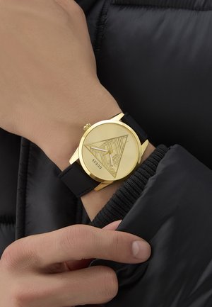 Ceas analog auriu cu un cadran cu model triunghiular și curea din silicon negru. Mâna poziționată aproape de mâneca unei jachete din puffer neagră.