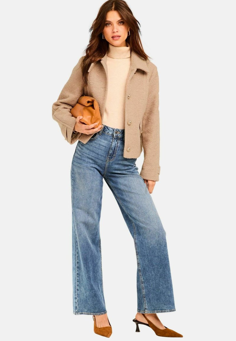 Giacca in misto lana di colore beige con bottoni a pressione, dolcevita avorio, jeans a vita alta di jeans azzurri a gamba larga e mules con tacco marroni.