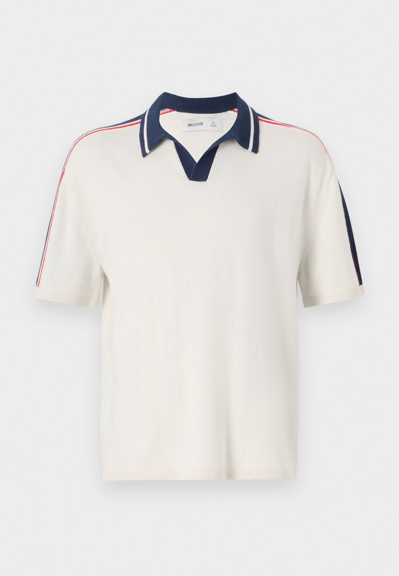Polo de manga corta blanco con cuello en V y color marino. Presenta franjas rojas y marinas en los hombros. Tejido de punto suave y texturizado.