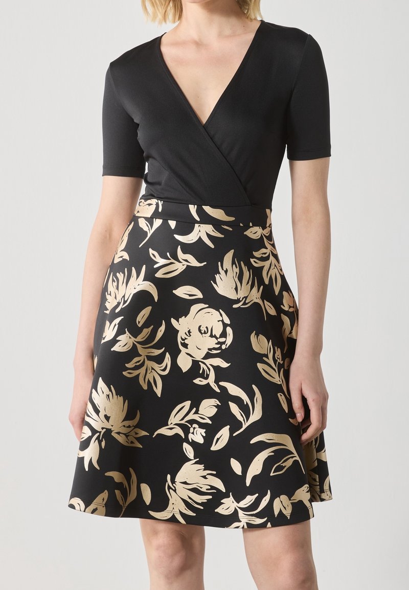 Femme portant un haut portefeuille noir à manches courtes et une jupe noire avec un imprimé floral beige, debout devant un fond uni.