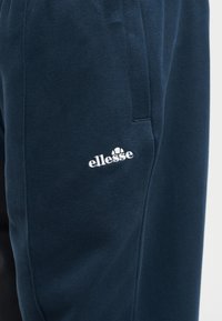 Μπλε σκούρο φούτερ παντελόνι με λευκό λογότυπο ellesse κεντημένο στον επάνω μηρό κοντά σε πλαϊνή τσέπη.