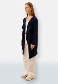 Marinblå lång cardigan med draperad front, ribbade ärmar och mjuk textur, tillsammans med krämfärgade vida byxor och beigefärgade sneakers.