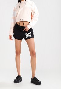 Hoodie cropped rosa chiaro con strisce bianche, coulisse regolabile. Pantaloncini neri con "IVY PARK" in bold bianco. Sneakers nere.