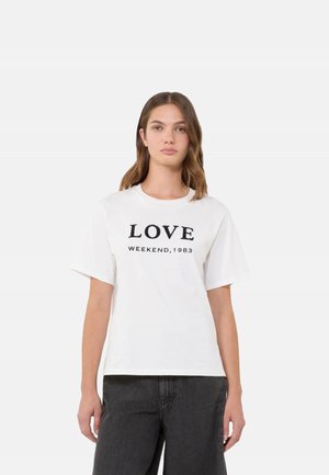 Giovane donna con capelli castani fino alle spalle, che indossa una maglietta bianca con la scritta "LOVE WEEKEND, 1983" e pantaloni neri, in posa su uno sfondo semplice.