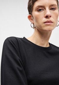 Μαύρη μπλούζα με μακριά μανίκια και στρογγυλή λαιμόκοψη, κατασκευασμένη από αδιάσπαστο ύφασμα, με ελαφρώς πεσμένους ώμους και εφαρμοστή γραμμή.