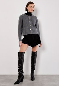 Frau steht an einer weißen Wand, trägt eine graue Knopffront-Strickjacke, ein schwarzes Rollkragenoberteil, schwarze Shorts mit Taschen und schwarze kniehohe Stiefel.