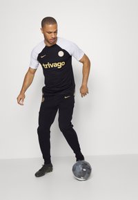 Tenue de sport en noir et blanc composée d'un t-shirt avec un logo et d'un pantalon de survêtement. La personne porte des chaussures de football noires avec des accents verts.