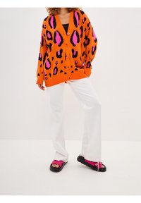 Oranžový kardigan s leopardím potiskem a černými a růžovými akcenty, s knoflíkovou přední částí, volný oversized střih, doplněný bílé kalhoty s širokými nohavicemi a růžové sandály.