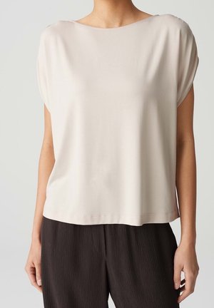 Femme portant un haut beige ample à manches courtes et un pantalon noir texturé à jambes larges sur un fond uni.