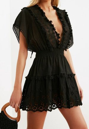 Kobieta w czarnej mini sukience z koronki i ażurowej tkaniny z głębokim dekoltem w serek i falbankowymi rękawami, trzymająca czarną plecioną torebkę z drewnianymi uchwytami.