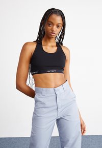 Black sports bra with a racerback design, featuring a branded elastic band. Paired with light gray, high-waisted pants. Smooth fabric texture.