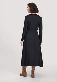 Schwarzes langärmliges Kleid mit taillierter Taille und fließendem Rock. Mit Knopfmanschetten und einer glatten Textur. Getragen mit kniebhigh braunen Stiefeln.