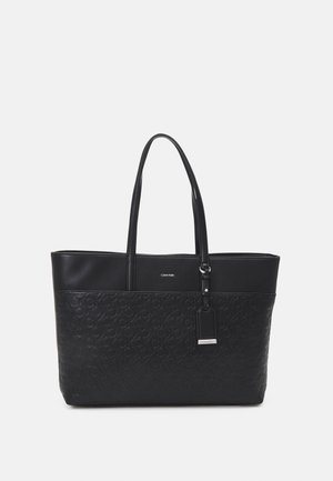 Czarna torba typu tote Calvin Klein z tłoczonym wzorem logo, dwoma paskami na ramię i zawieszką z srebrnym detalem na jednolitym tle.