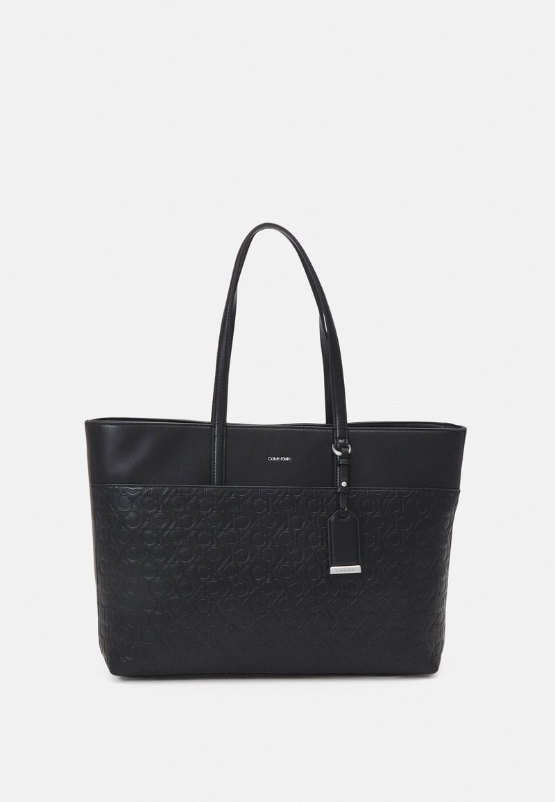 Sac cabas noir Calvin Klein avec motif de logo en relief, deux bretelles d'épaule et une étiquette suspendue avec détail argenté sur fond uni.