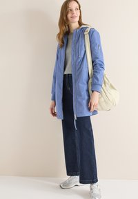 Femme portant une veste à capuche bleue, un t-shirt gris, un jean large foncé, des baskets argentées, et portant un grand sac à bandoulière beige.