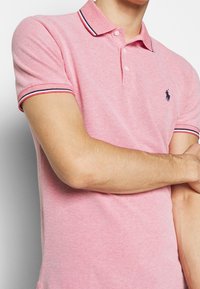 Polo shirt van roze stof met een gestructureerde weef. Heeft een kraag en knoopsluiting, versierd met marineblauwe en rode strepen op de mouwen.