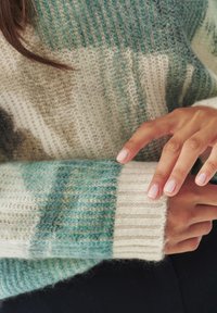 Hände mit gepflegten Nägeln, die den Ärmel eines gemütlichen Strickpullovers in sanften Blau-, Beige- und Cremetönen anpassen.