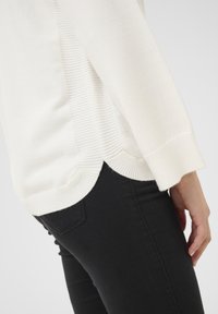 Vit tröja med ribbade sidopaneler och rundad fåll, kombinerad med svarta jeans. Mjuk textur, lös passform och ärmar i halv längd.