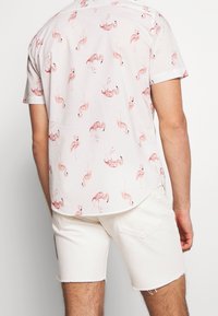 Camicia a maniche corte di colore rosa chiaro con motivo fenicottero, dotata di colletto con bottoni e orlo arrotondato, abbinata a pantaloni shorts in denim bianchi.