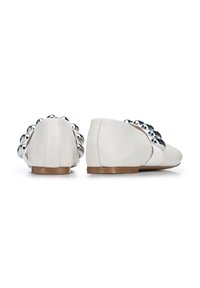 Zapatos de cuero blanco sin cordones con punteras redondeadas, que presentan una fila decorativa de bolas de cromo en la parte trasera y una suela marrón.