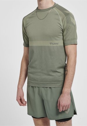 Mand iført olivengrønt hummel sportstøjssæt med kortærmet skjorte og shorts, kædehalskæde og tatoveringer på højre underarm og lår.