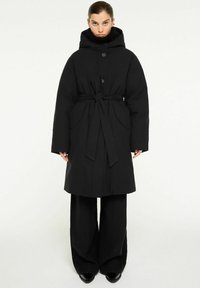 Svart vadderad jacka med hög krage, midjeband och stora sidofickor. Har en huva och knäppning med knappar. Krossad med vida svarta byxor.