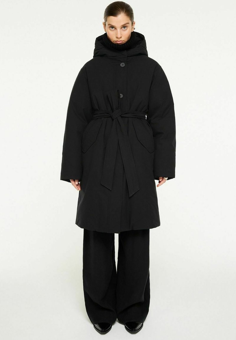 Svart vadderad jacka med hög krage, midjeband och stora sidofickor. Har en huva och knäppning med knappar. Krossad med vida svarta byxor.