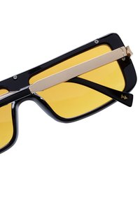Zwarte zonnebril met opvallende rechthoekige monturen, gele glazen en een metallic gouden accentbalk bovenaan. Geprofileerde pootjes voor extra grip.