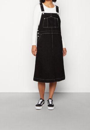 Robe salopette en jean noir avec surpiqûres blanches, poches latérales et bretelles ajustables. Associée à des baskets noires et blanches.