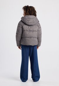 Chaqueta tipo abrigo en un patrón de cuadros marrón y gris, con una gran capucha y un acolchado grueso, combinada con pantalones de mezclilla de pierna ancha.