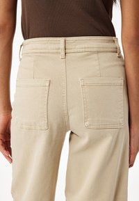Beige high-waisted broek met een rechte pijp, voorzien van twee achterzakken, gemaakt van een gestructureerde stof. De tailleband heeft riemlussen.