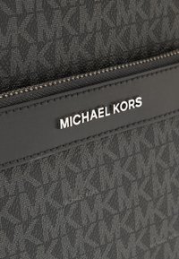 Matériau synthétique noir à motif avec une finition texturée. Équipé d'une fermeture éclair en métal argenté et d'une plaque rectangulaire embossée avec "MICHAEL KORS."