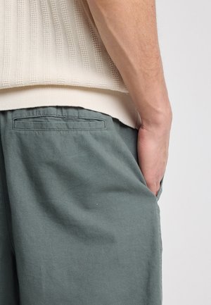 Man wearing cream knit sweater and gray pants with hand in side pocket, shown from waist to mid-thigh against plain background.