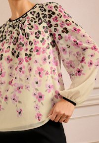 Blusa trasparente con una base crema, decorata con motivi floreali rosa e neri e pattern leopardati, maniche lunghe a sbuffo e rifinitura del collo nera.