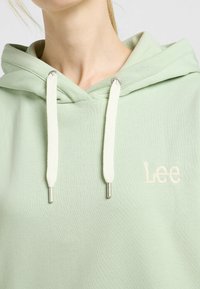 Sudadera de color verde claro con una textura suave, que presenta un bolsillo delantero, cordones blancos con extremos plateados y "Lee" bordado en blanco.