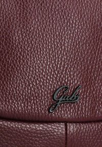 Close-up van bordeauxkleurig textuurleer met zwart "Gabs" logo-embleem op het oppervlak.