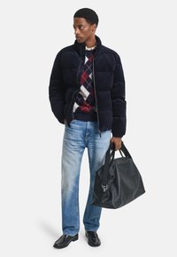 Marineblauer Pufferjacke, gestepptes Design, mit Reißverschluss. Roter, weißer und blauer Argyle-Pullover darunter, hellblaue Jeans, schwarze Lederschuhe, schwarze Tasche.