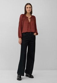 Roestkleurige blouse met een keyhole-halslijn, geplooide details en lange mouwen, gecombineerd met hoge zwarte wijde jeans en glanzende schoenen.