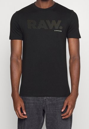 Mężczyzna ubrany w czarną koszulkę z krótkim rękawem z napisem "RAW." w stylizowanej, ukośnej linii oraz małym logo "G-STAR RAW" na piersi, połączoną z ciemnymi dżinsami.