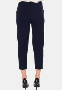 Vista posteriore di una persona che indossa pantaloni capri blu scuro e scarpe con tacco nere, in piedi contro uno sfondo neutro.